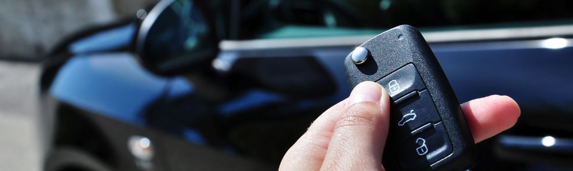 a left hand holds a black key fob and in the background is a black vehicle parked on the road