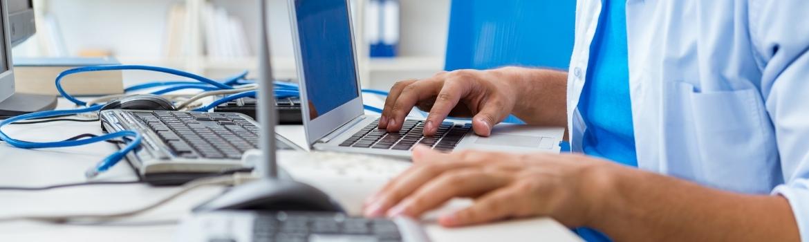 a person types on a laptop keyboard with their right hand while their left hand rests on the edge of a desk.  Behind the laptop is a keyboard with a blue ethernet cable on top of the keyboard not plugged into anything.