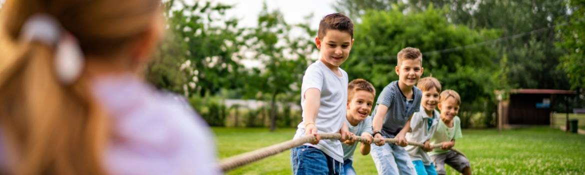 in the foreground, a girl with a pony tail is pulling on a rope of tug-a-war and in the background are 5 young boys, around seven to nine years old pulling on the other end of the rope as this game takes place on a big grass area