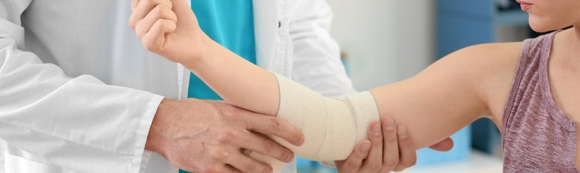 a doctor wraps a white bandage over the elbow of a girl's right warm, who appears to be young and wearing a maroon tank top