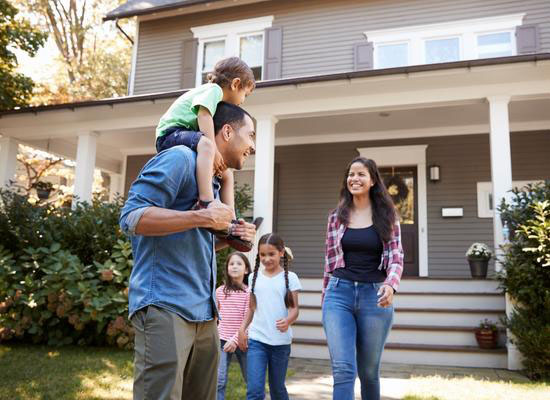 A child rides on dads shoulders with mom and two young daughters walk away from their two story home toward the sidewalk